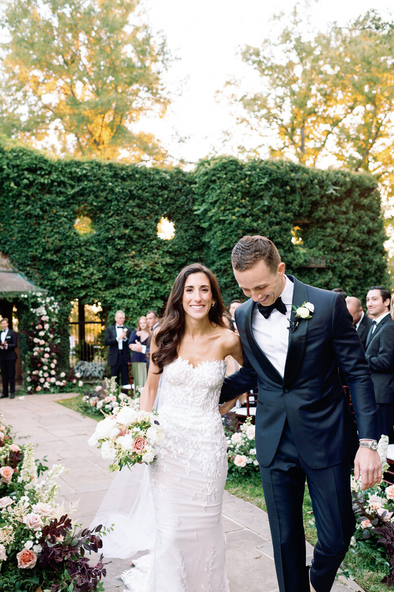 Newlyweds walking down aisle after ceremony in garden venue with floral-lined pathway and ivy-covered hedge backdrop