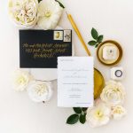 Elegant wedding stationery flat lay featuring black envelope with gold calligraphy, white invitation, and cream florals
