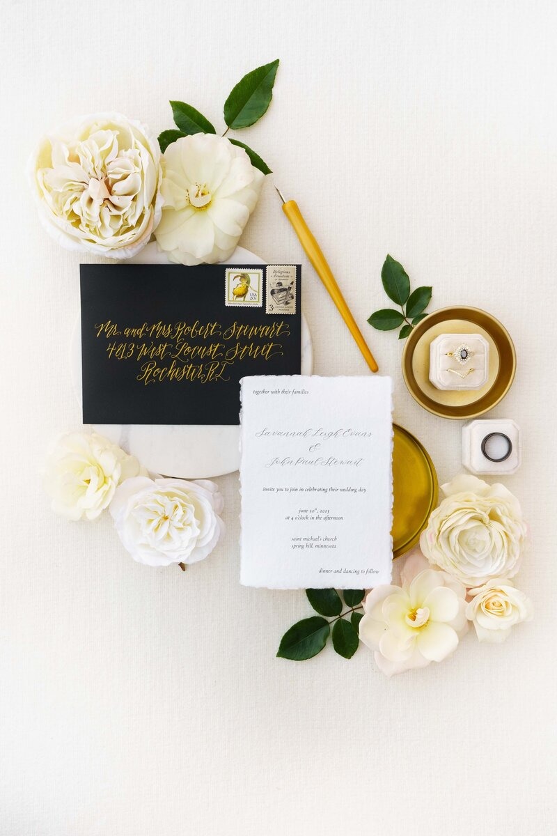 Elegant wedding stationery flat lay featuring black envelope with gold calligraphy, white invitation, and cream florals