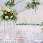 Tall tropical wedding centerpieces with monstera leaves, roses, and hydrangeas on clear stands at reception tables