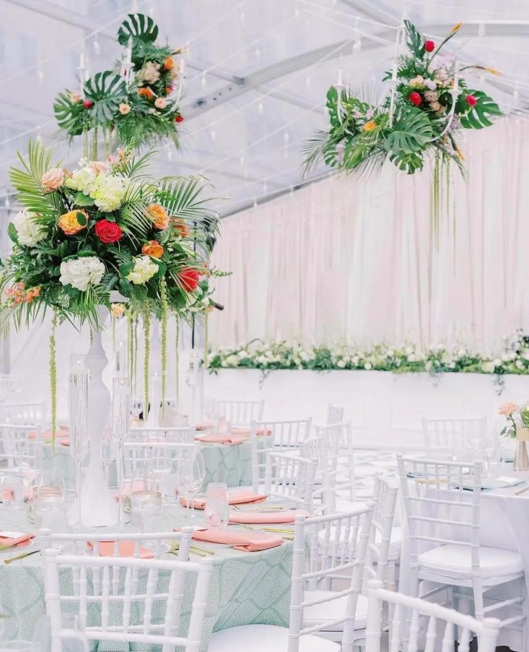 Tall tropical wedding centerpieces with monstera leaves, roses, and hydrangeas on clear stands at reception tables