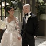 Smiling bride in long-sleeve lace gown with white bouquet walking hand-in-hand with groom in black tuxedo