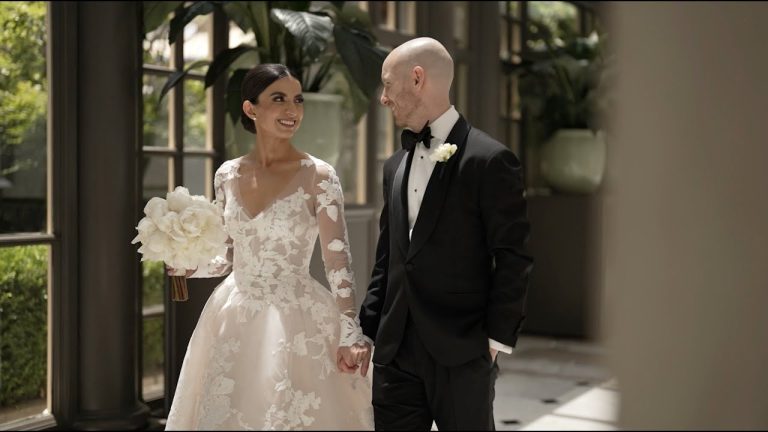 Smiling bride in long-sleeve lace gown with white bouquet walking hand-in-hand with groom in black tuxedo