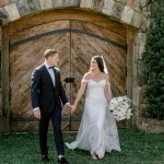 Bride and groom holding hands in front of rustic wooden barn door with stone archway