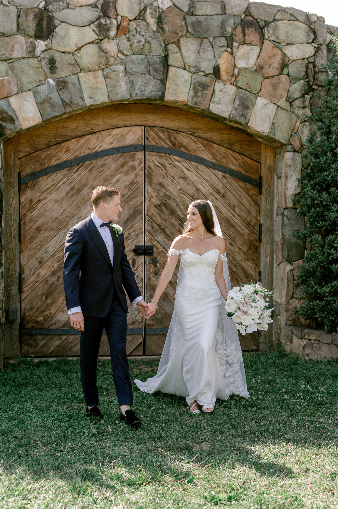 Bride and groom holding hands in front of rustic wooden barn door with stone archway