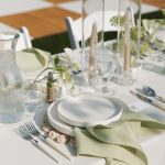 Modern outdoor wedding table setting with white plates, sage green napkins, tall taper candles, and delicate floral centerpieces