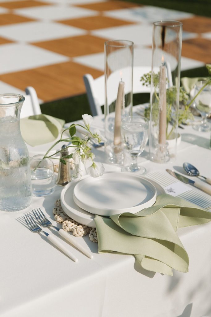 Modern outdoor wedding table setting with white plates, sage green napkins, tall taper candles, and delicate floral centerpieces