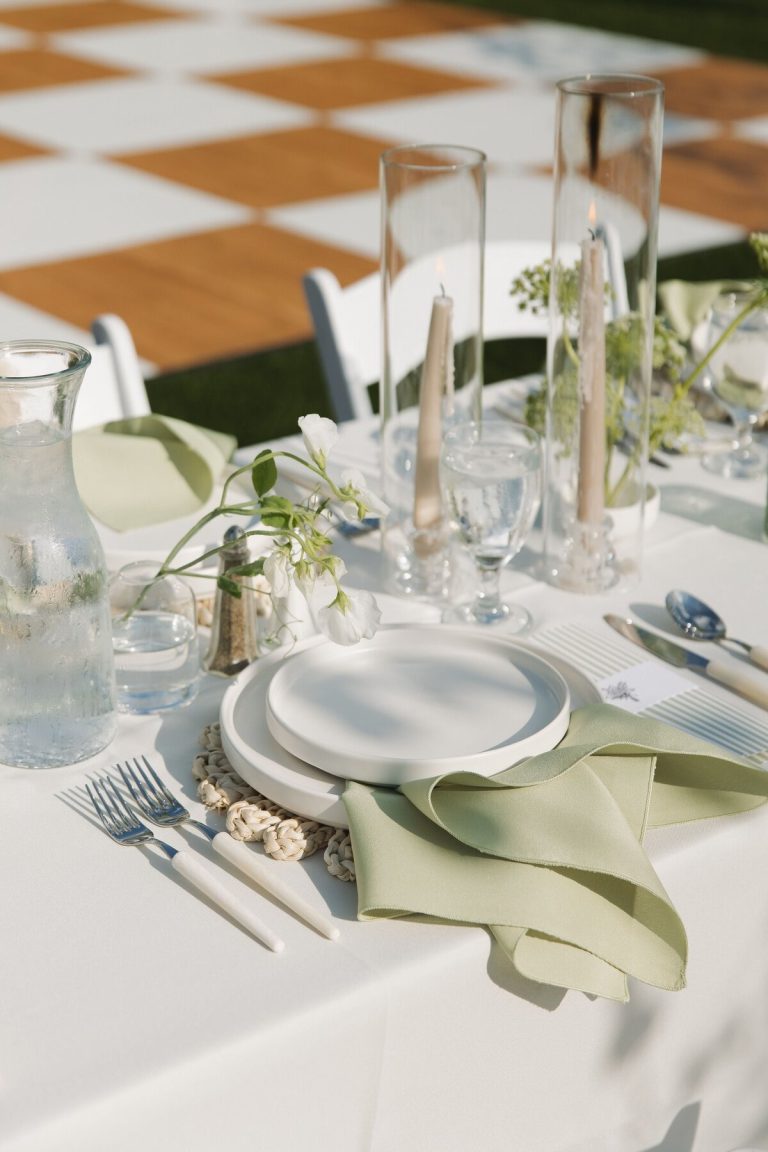 Modern outdoor wedding table setting with white plates, sage green napkins, tall taper candles, and delicate floral centerpieces