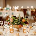Elegant wedding centerpiece with peach roses, burgundy dahlias, and greenery in gold pedestal vase surrounded by amber votives