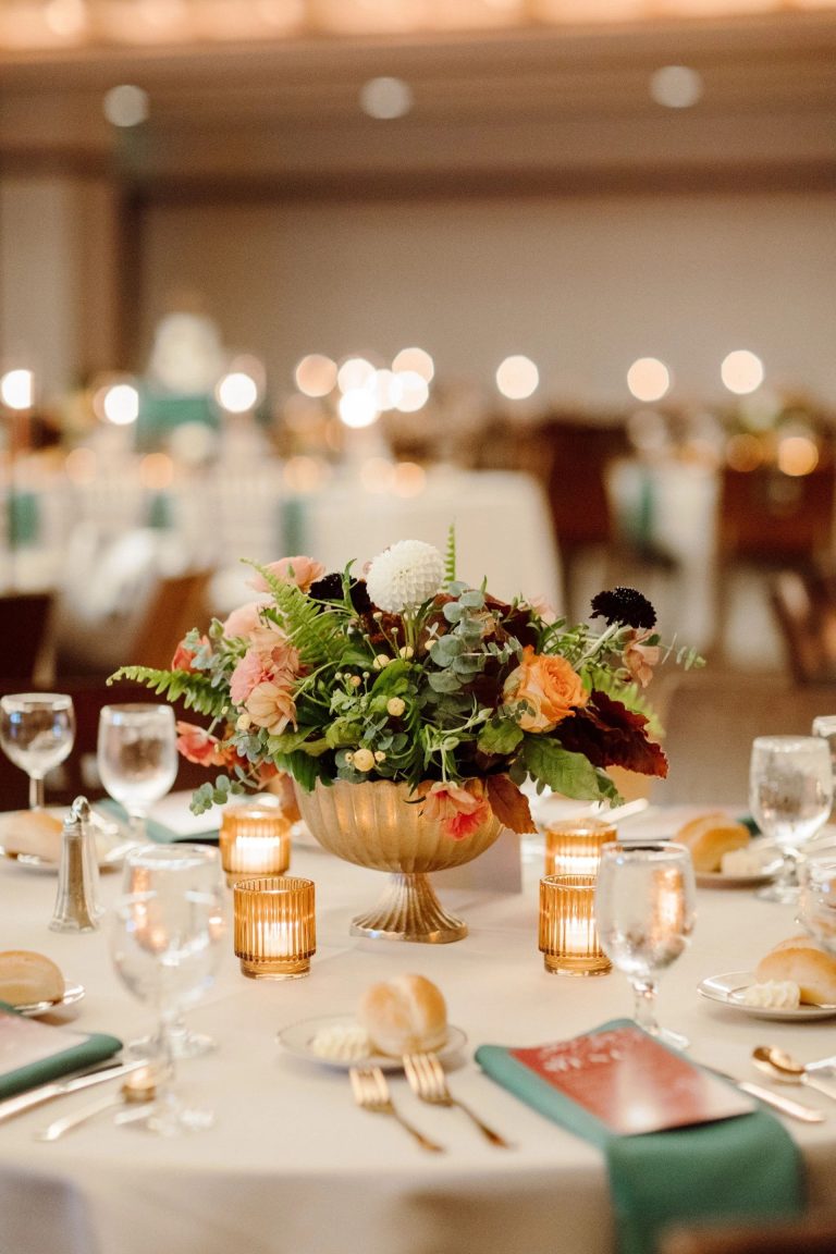Elegant wedding centerpiece with peach roses, burgundy dahlias, and greenery in gold pedestal vase surrounded by amber votives