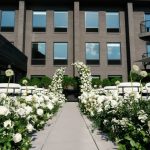 Outdoor wedding ceremony setup with white chairs and lush white floral arrangements lining a center aisle between modern buildings