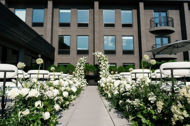 Outdoor wedding ceremony setup with white chairs and lush white floral arrangements lining a center aisle between modern buildings