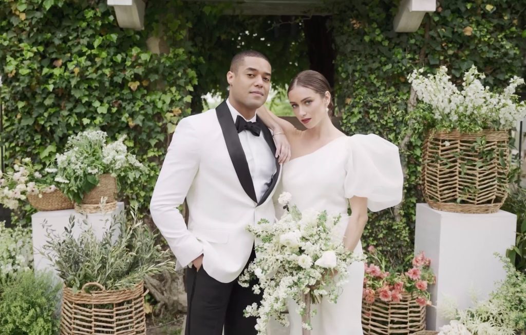 Groom in white tuxedo jacket and bride in elegant puff-sleeve gown holding white bouquet against ivy-covered wall with wicker basket decor