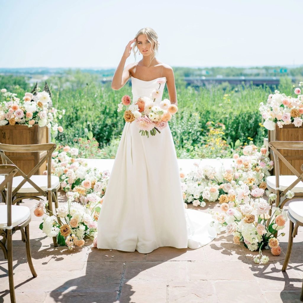 Bride in strapless white gown holding pastel bouquet at outdoor ceremony with lush floral arrangements and vineyard views