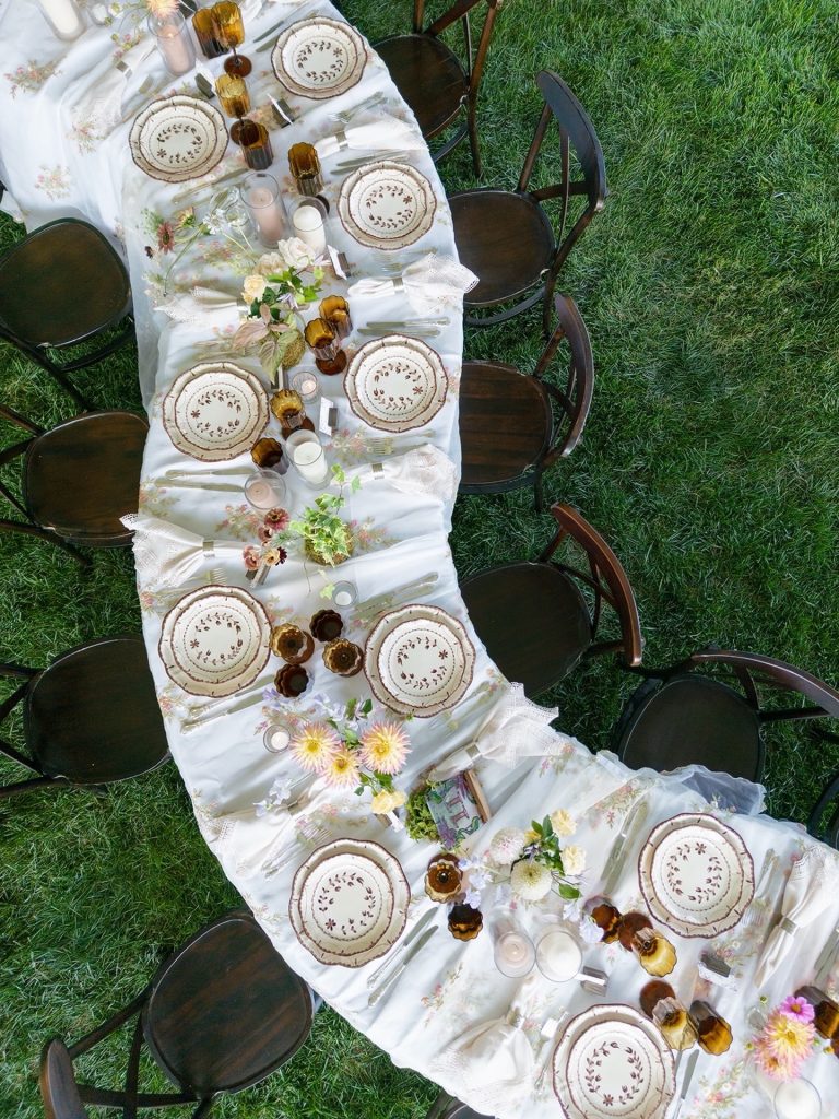 Aerial view of garden party table with decorative plates, amber glasses, and floral centerpieces on white tablecloth
