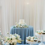 White tiered wedding cake with blue hydrangea florals on dusty blue tablecloth surrounded by cupcakes
