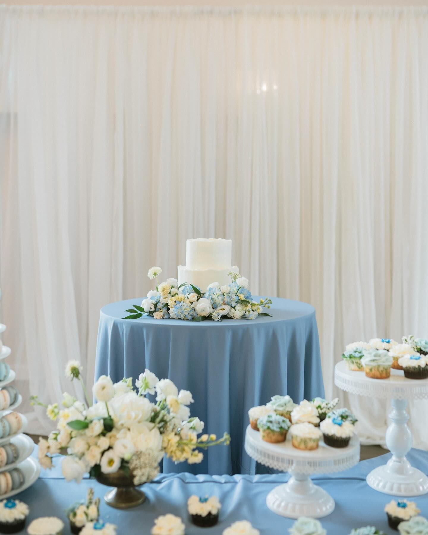 White tiered wedding cake with blue hydrangea florals on dusty blue tablecloth surrounded by cupcakes