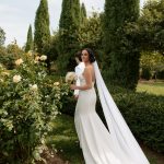 Bride and groom in garden with yellow rose bushes and tall cypress trees, her cathedral veil flowing behind