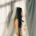 Bride in profile wearing long veil and strapless lace gown, framed by soft curtains