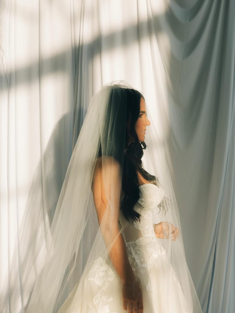 Bride in profile wearing long veil and strapless lace gown, framed by soft curtains