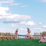 Waterfront ceremony site featuring circular floral arch in pink and purple hues with wooden cross-back chairs arranged on green lawn