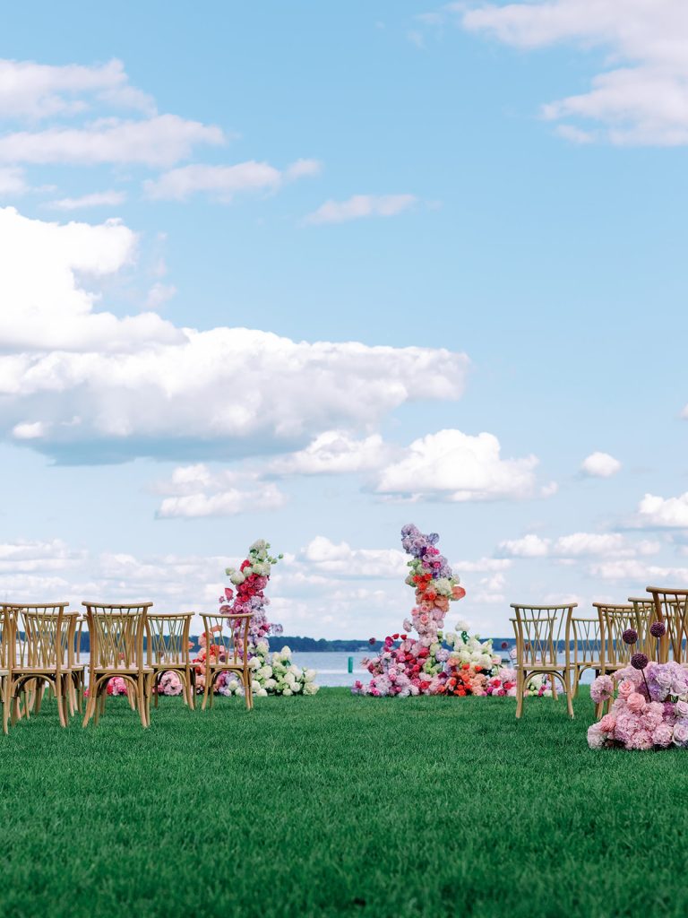 Waterfront ceremony site featuring circular floral arch in pink and purple hues with wooden cross-back chairs arranged on green lawn