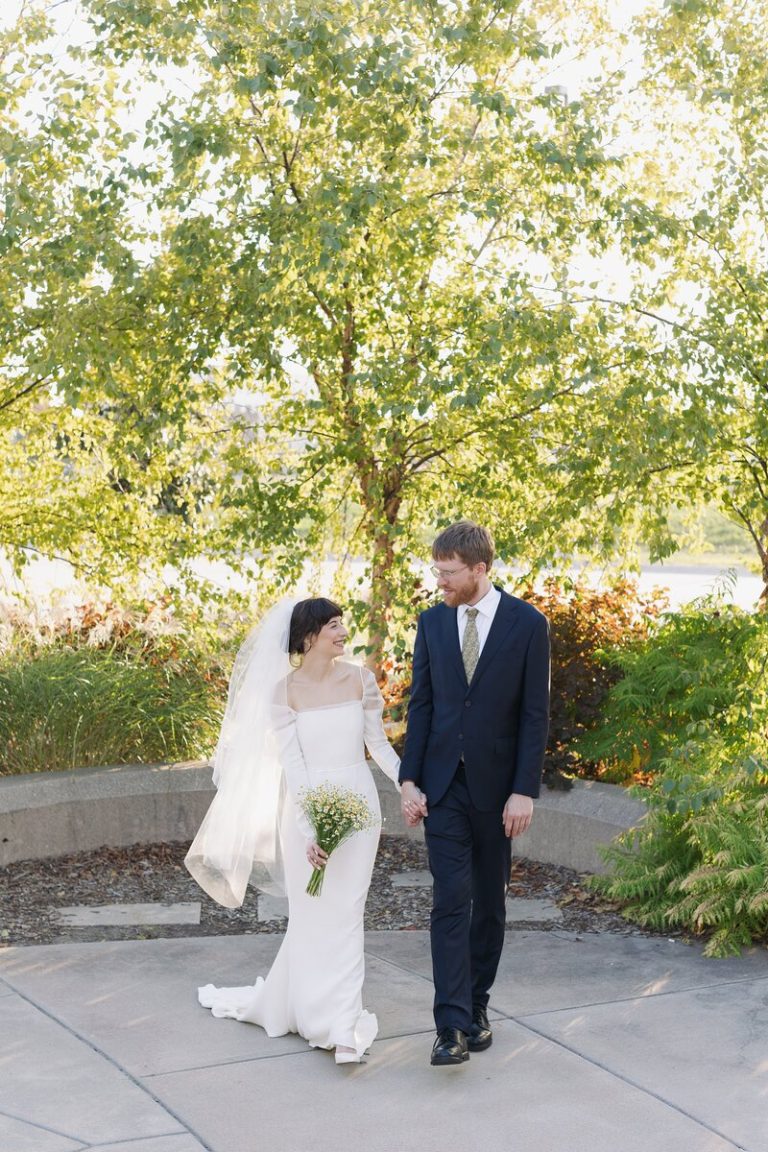 Bride and groom smiling at each other while holding hands outdoors with baby's breath bouquet