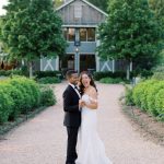 Bride and groom embracing on path in front of estate house surrounded by lush greenery