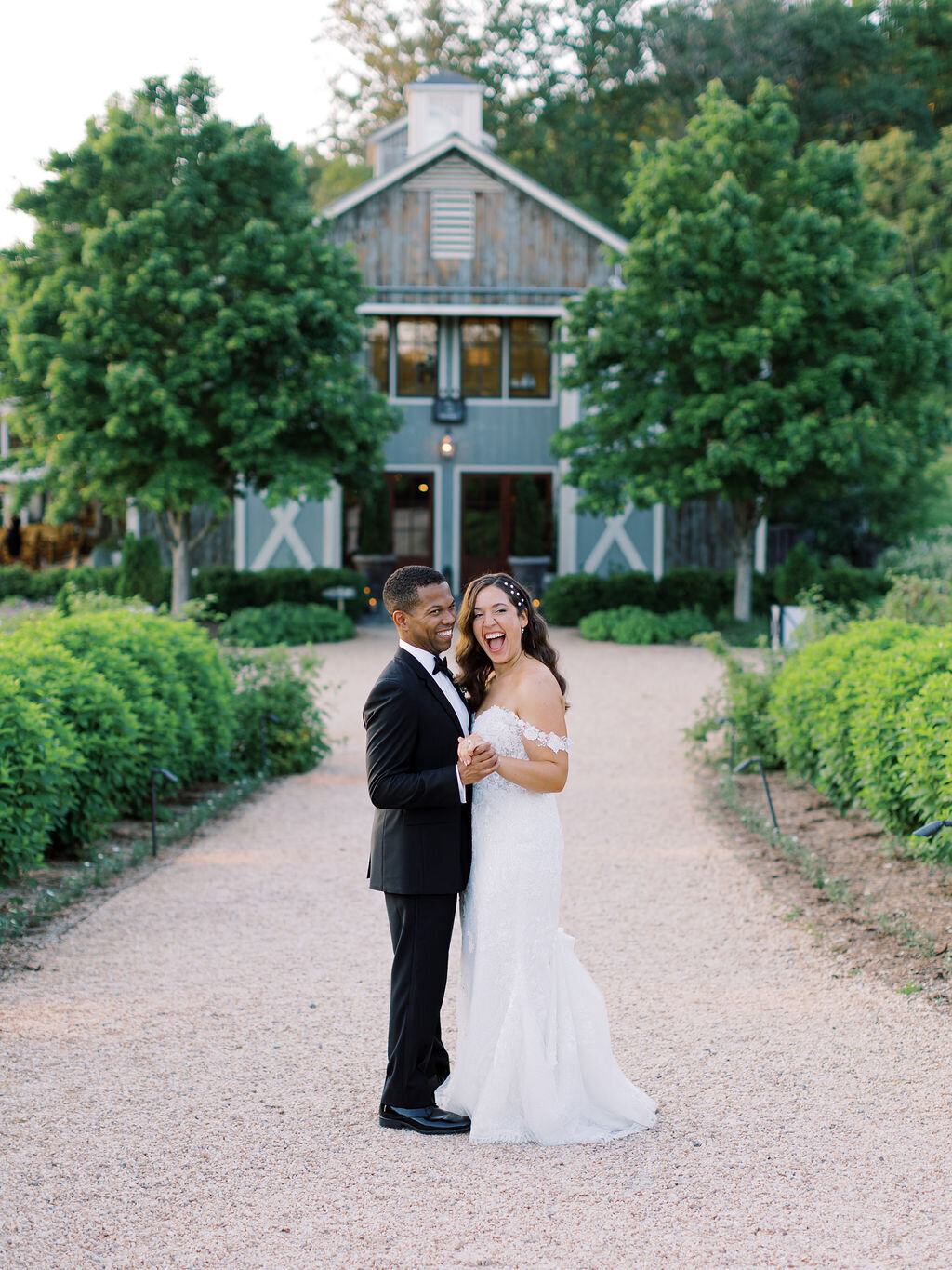Bride and groom embracing on path in front of estate house surrounded by lush greenery