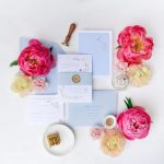 Flat lay of wedding invitation suite in soft blue with pink peonies, ranunculus, wax seal, and ring in gold box