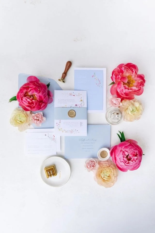 Flat lay of wedding invitation suite in soft blue with pink peonies, ranunculus, wax seal, and ring in gold box