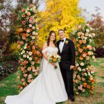 Bride in off-shoulder ball gown and groom in black tuxedo framed by autumn floral arrangements outdoors