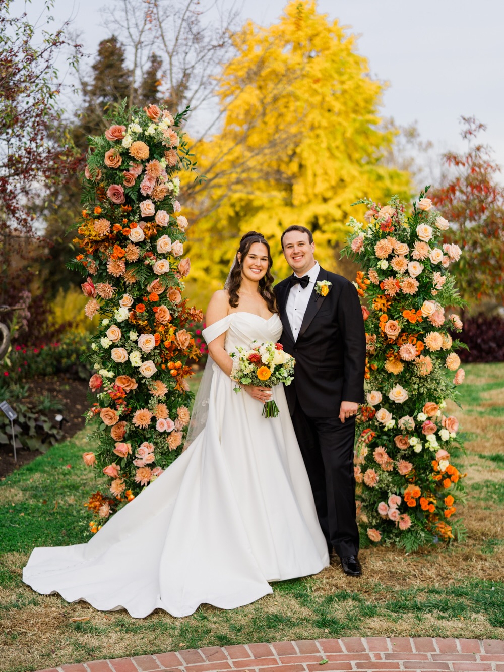 Bride in off-shoulder ball gown and groom in black tuxedo framed by autumn floral arrangements outdoors