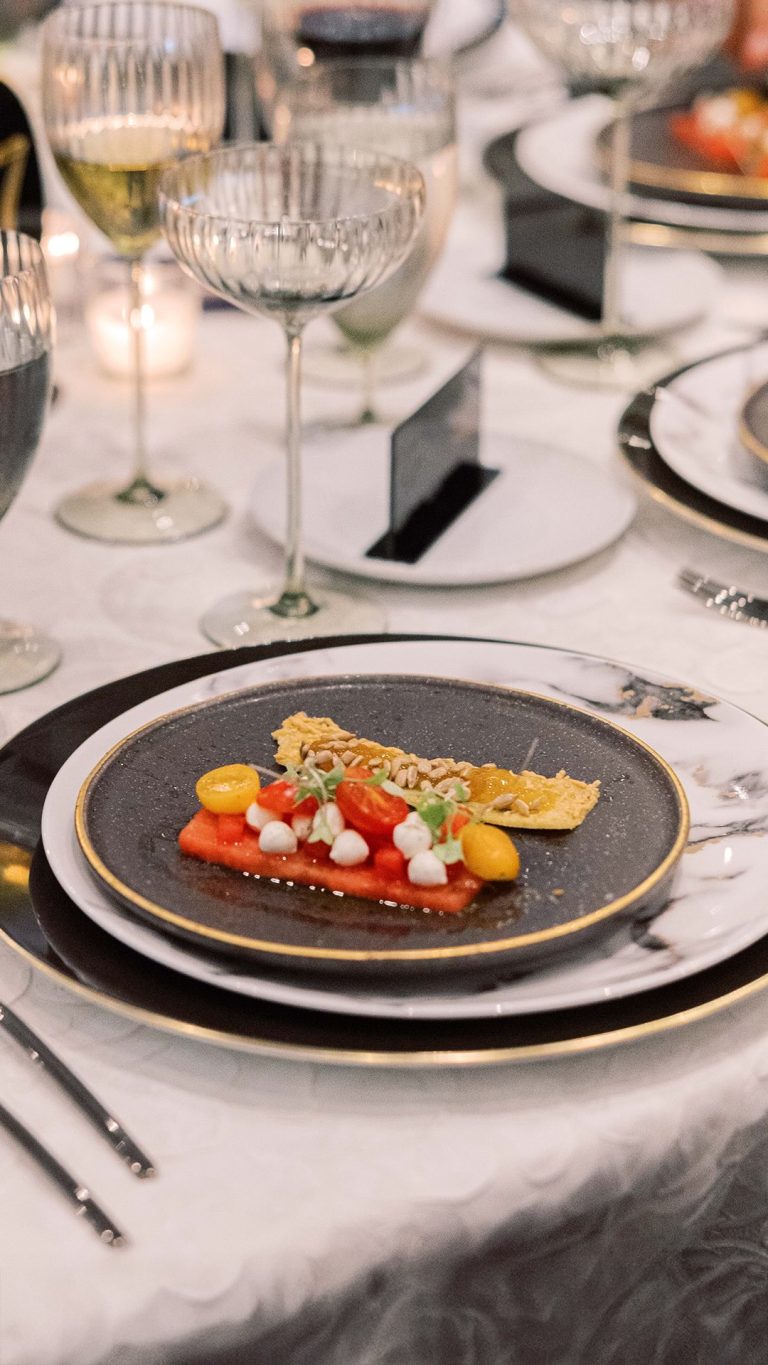 Fine dining appetizer with watermelon, cherry tomatoes, white cheese dots, and crispy tuile on gold-rimmed dark plate