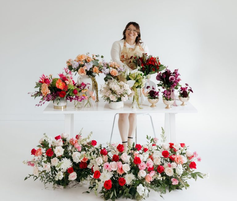 Bride in blush dress standing behind elaborate floral arrangements in burgundy, pink, and white