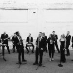 Professional wedding band in black formal attire performing with instruments against white brick wall
