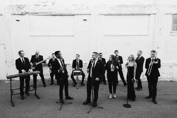 Professional wedding band in black formal attire performing with instruments against white brick wall