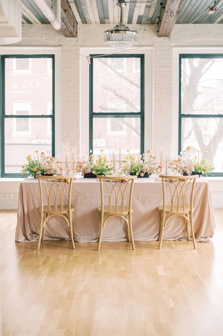 Wedding reception table featuring bentwood chairs, blush linens, floral arrangements, and acrylic candlesticks in white brick venue