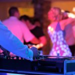 DJ in blue suit mixing music at turntables while wedding guests dance under pink and blue lighting