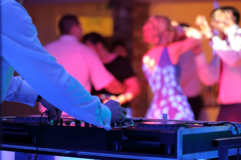 DJ in blue suit mixing music at turntables while wedding guests dance under pink and blue lighting