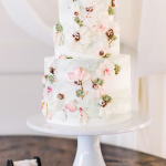 Three-tier white wedding cake adorned with pink and white sugar flowers and greenery on a white pedestal stand