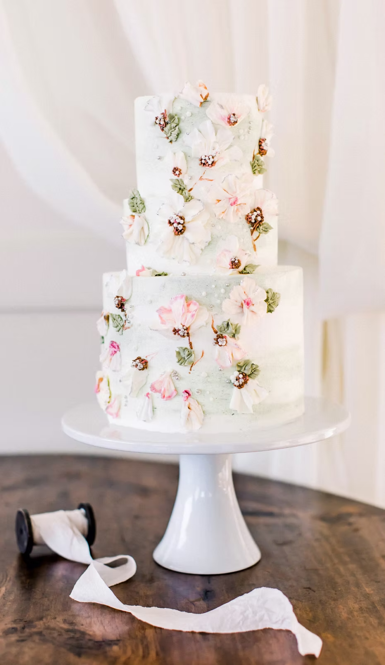 Three-tier white wedding cake adorned with pink and white sugar flowers and greenery on a white pedestal stand