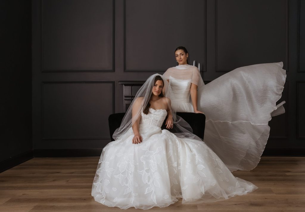 Two brides modeling wedding gowns in an elegant studio with dark walls, one seated in a ballgown with veil, the other standing in a flowing tulle dress