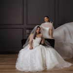 Two brides modeling wedding gowns in an elegant studio with dark walls, one seated in a ballgown with veil, the other standing in a flowing tulle dress