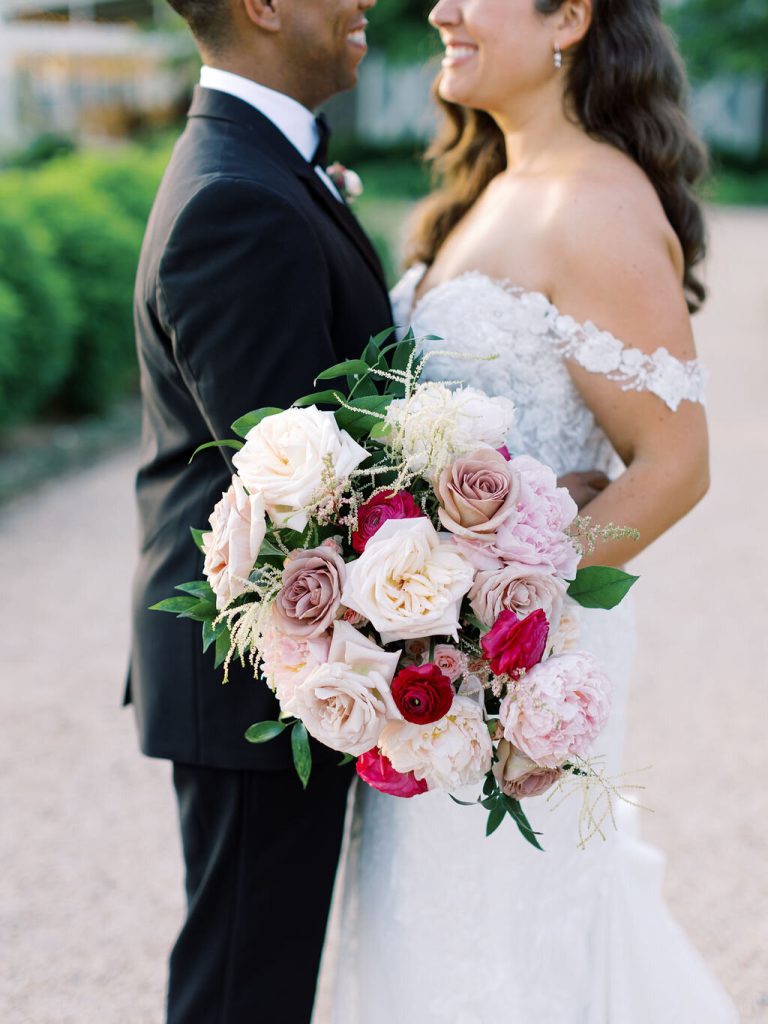 Bride and groom standing together, showcasing elegant bridal bouquet with roses, peonies, and greenery in pink and burgundy palette