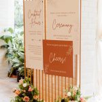 Wooden wedding signage display with ceremony and cocktail hour details adorned with orange roses and mixed floral arrangements