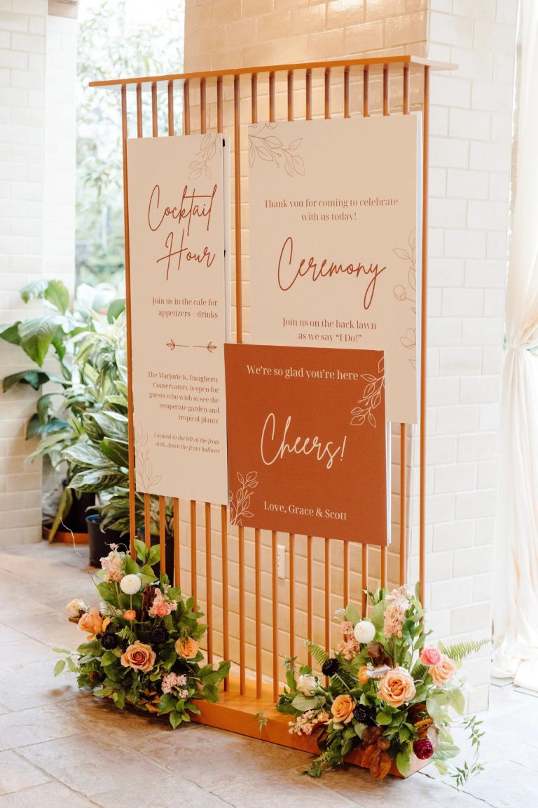 Wooden wedding signage display with ceremony and cocktail hour details adorned with orange roses and mixed floral arrangements