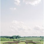 Wooden bench seating arranged in rows on a green lawn facing rolling vineyard fields under a cloudy sky