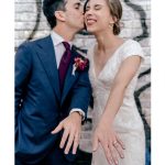Newlyweds displaying their wedding rings, groom in navy suit kissing smiling bride's cheek