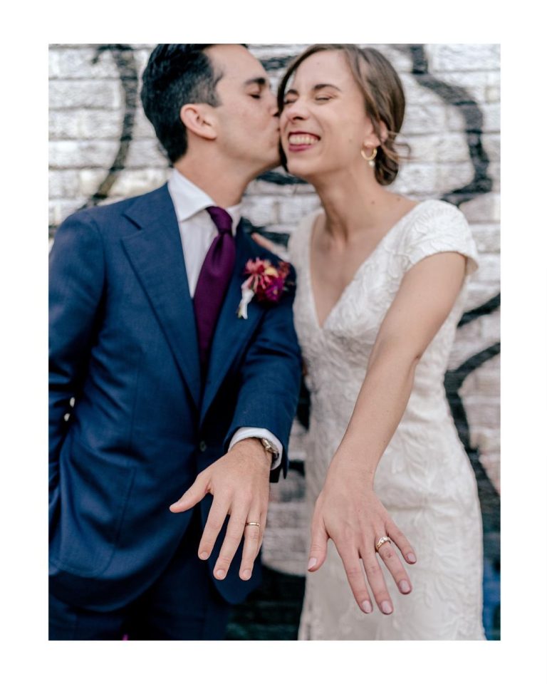 Newlyweds displaying their wedding rings, groom in navy suit kissing smiling bride's cheek